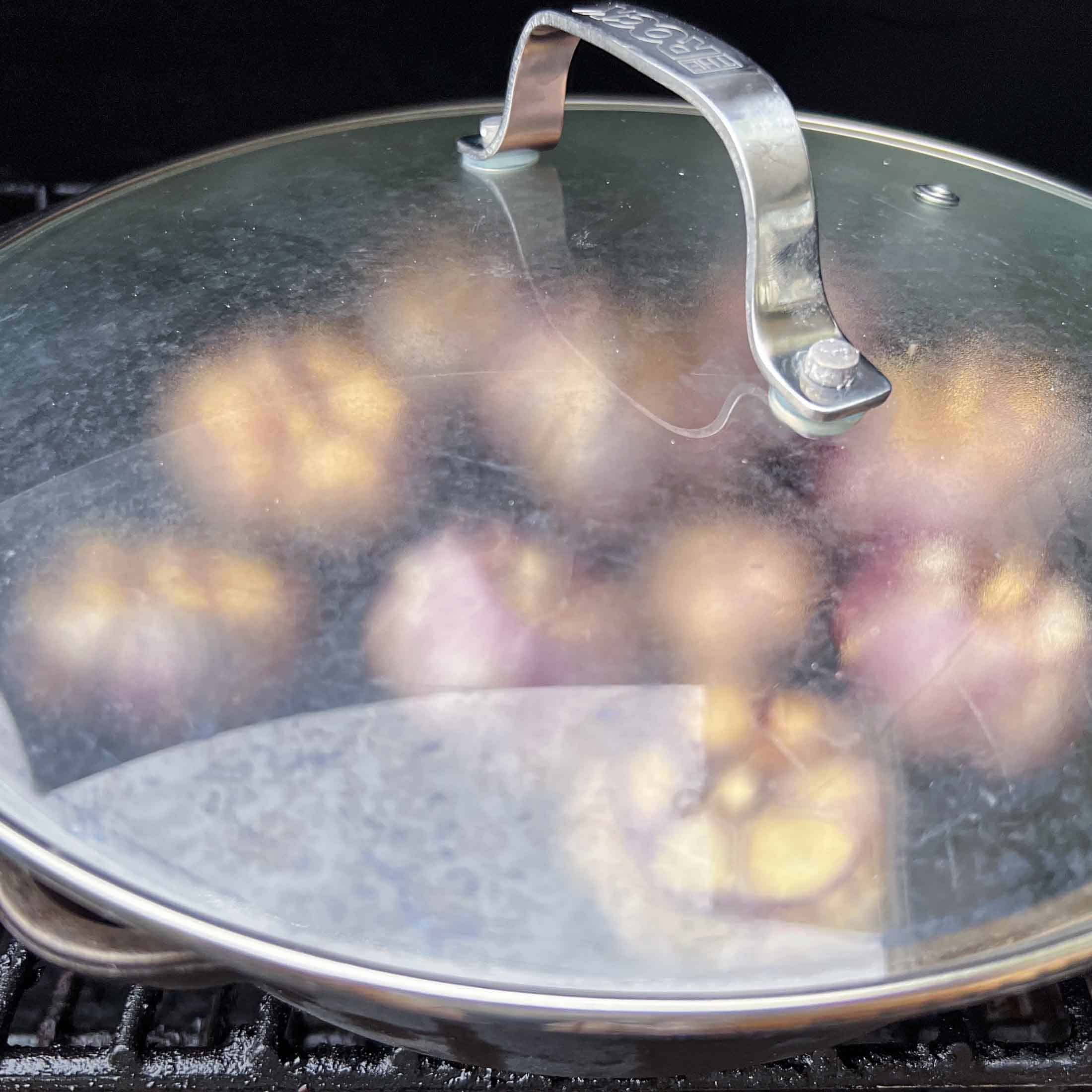 Garlic in a cast iron pan covered by a glass lid on a pellet grill.