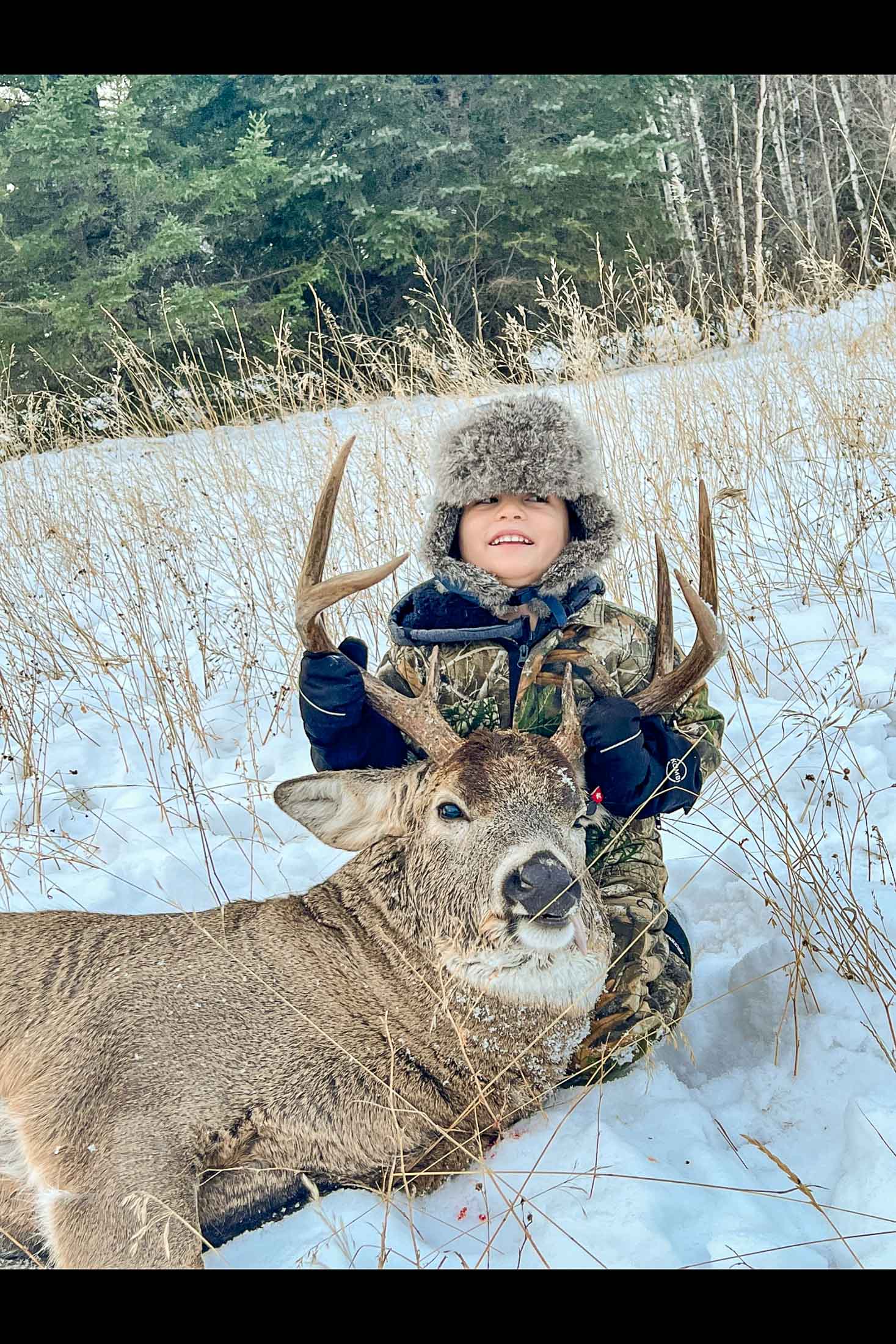 A young boy holding the antlers of a freshly harvested white tail buck in the snow.