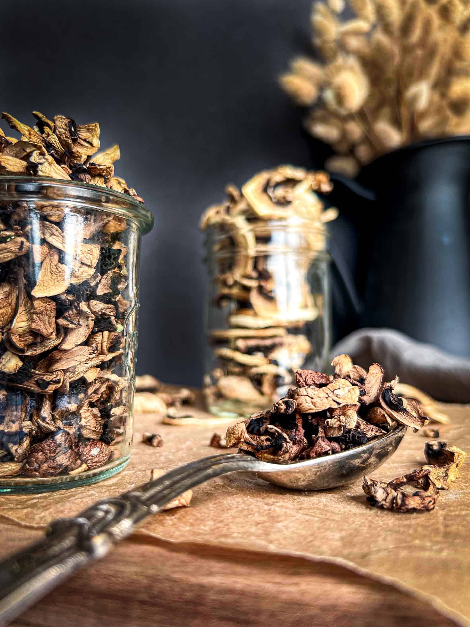 A closeup view of dehydrated mushroom pieces on a silver 1 tablespoon measuring spoon with jarred dried mushrooms in the foreground.