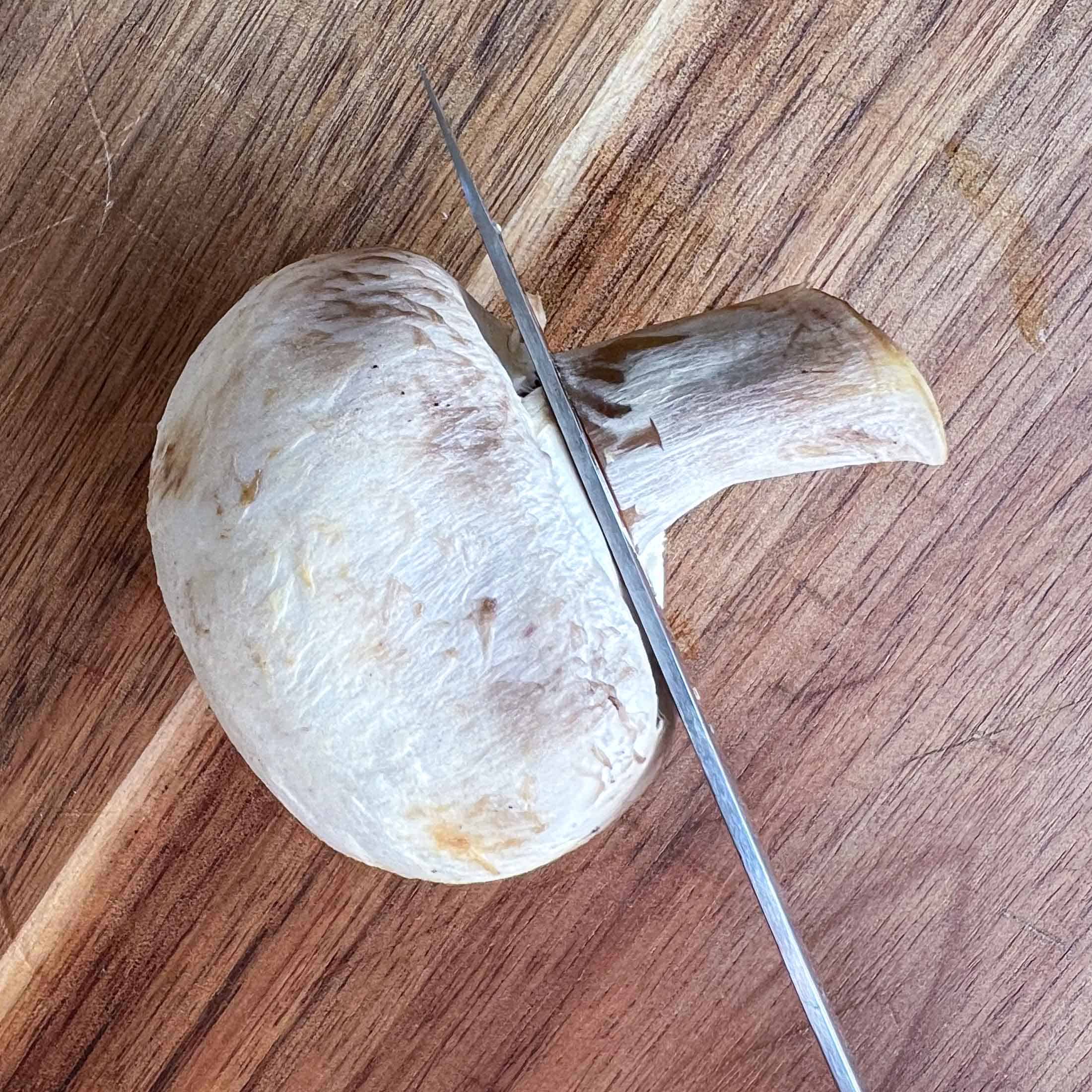 A sharp knife cutting the stem of a white mushroom off.