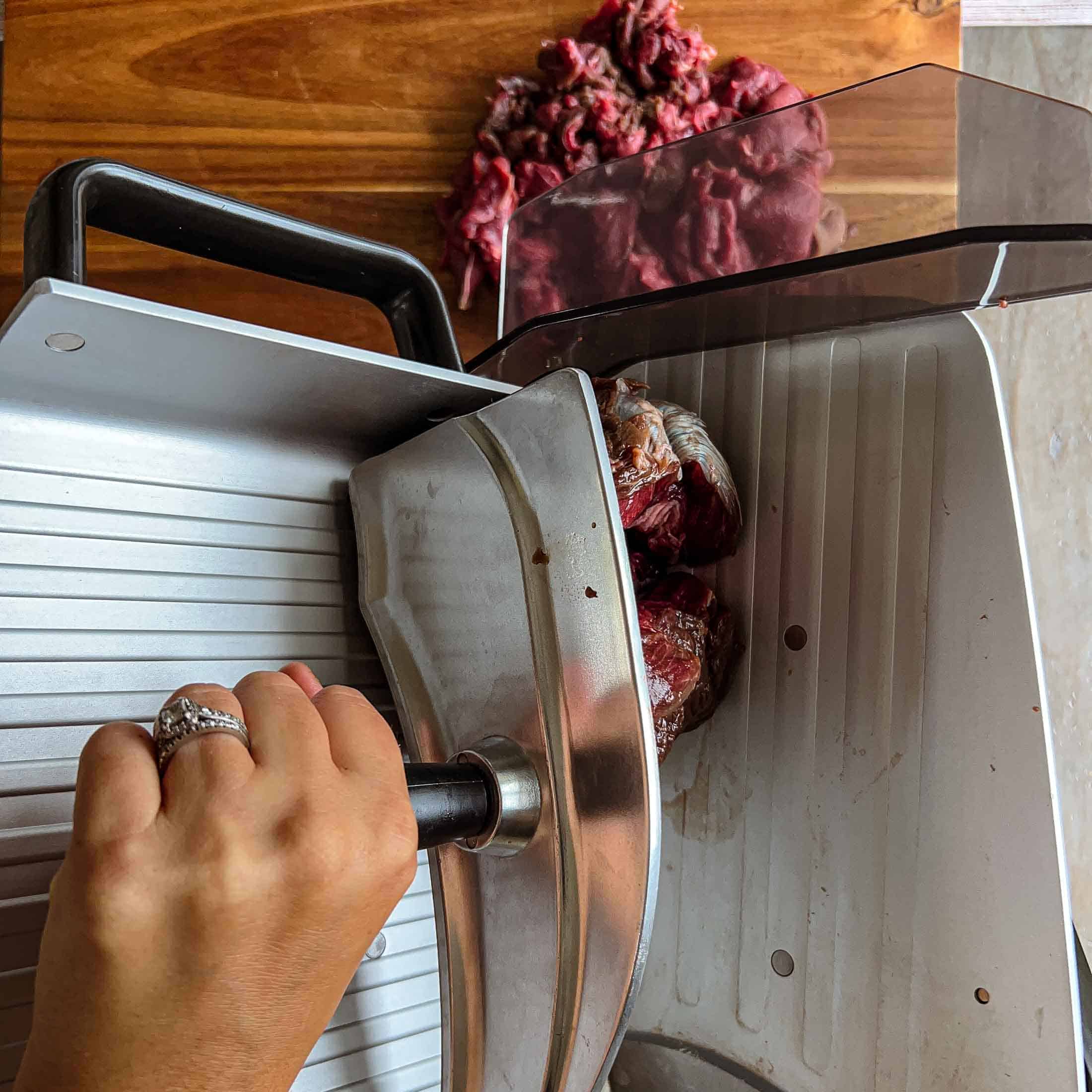 Raw venison roast being pushed through a meat slicer.