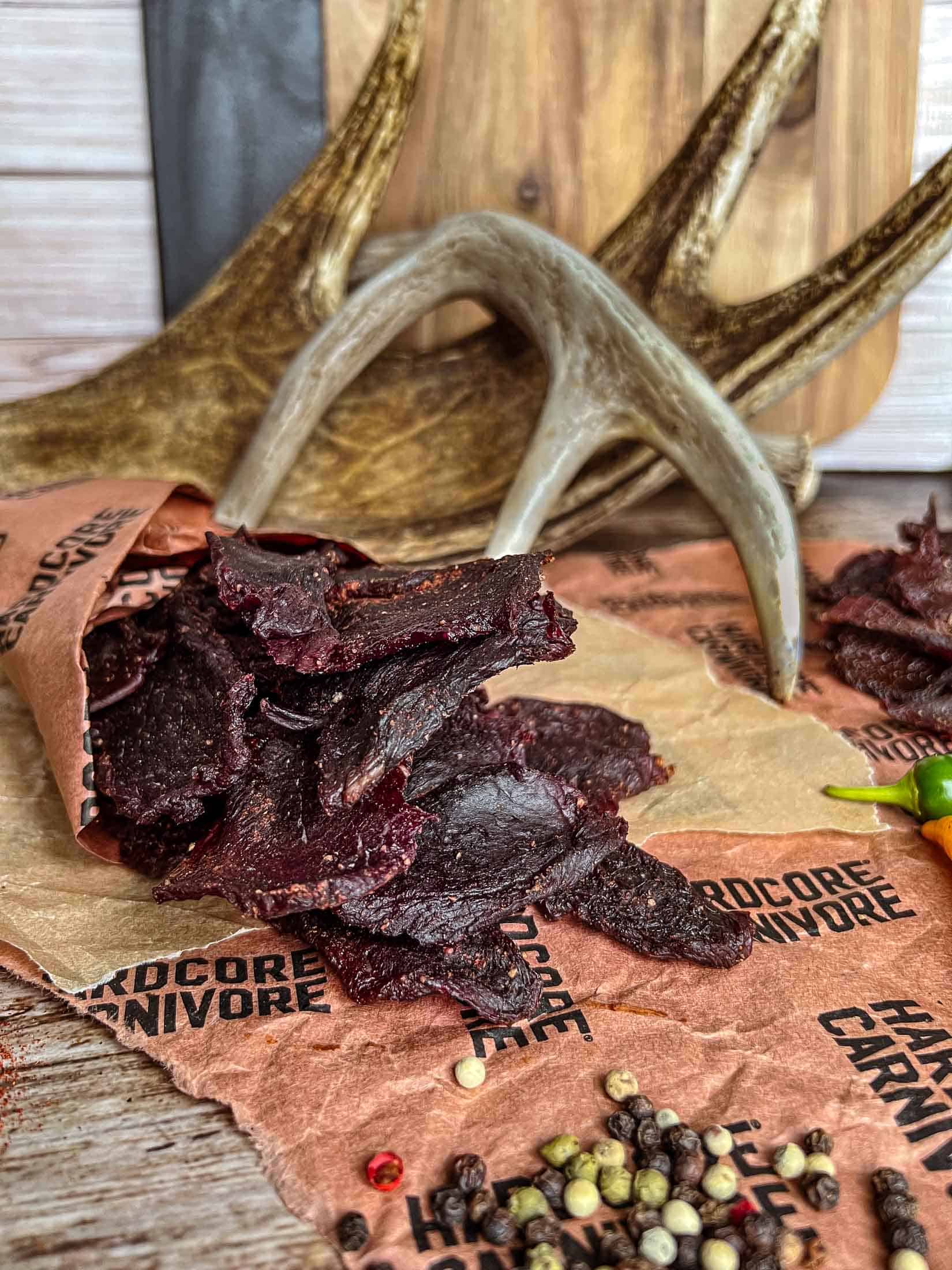 Venison jerky spilling out of a butcher paper cone with full peppercorns around and deer antlers behind.