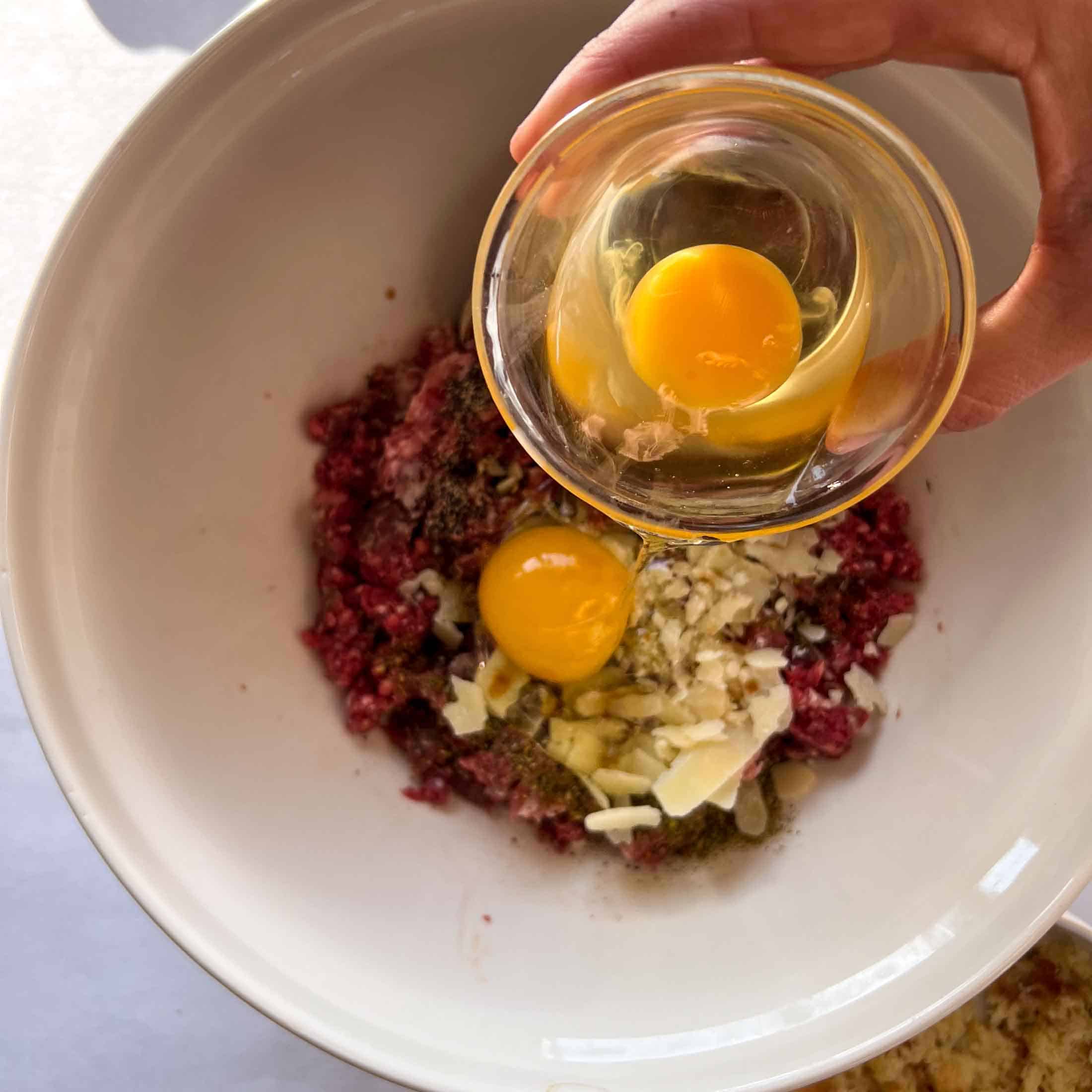 Two eggs being dumped into a cream bowl filled with raw venison, raw pork, and seasonings.
