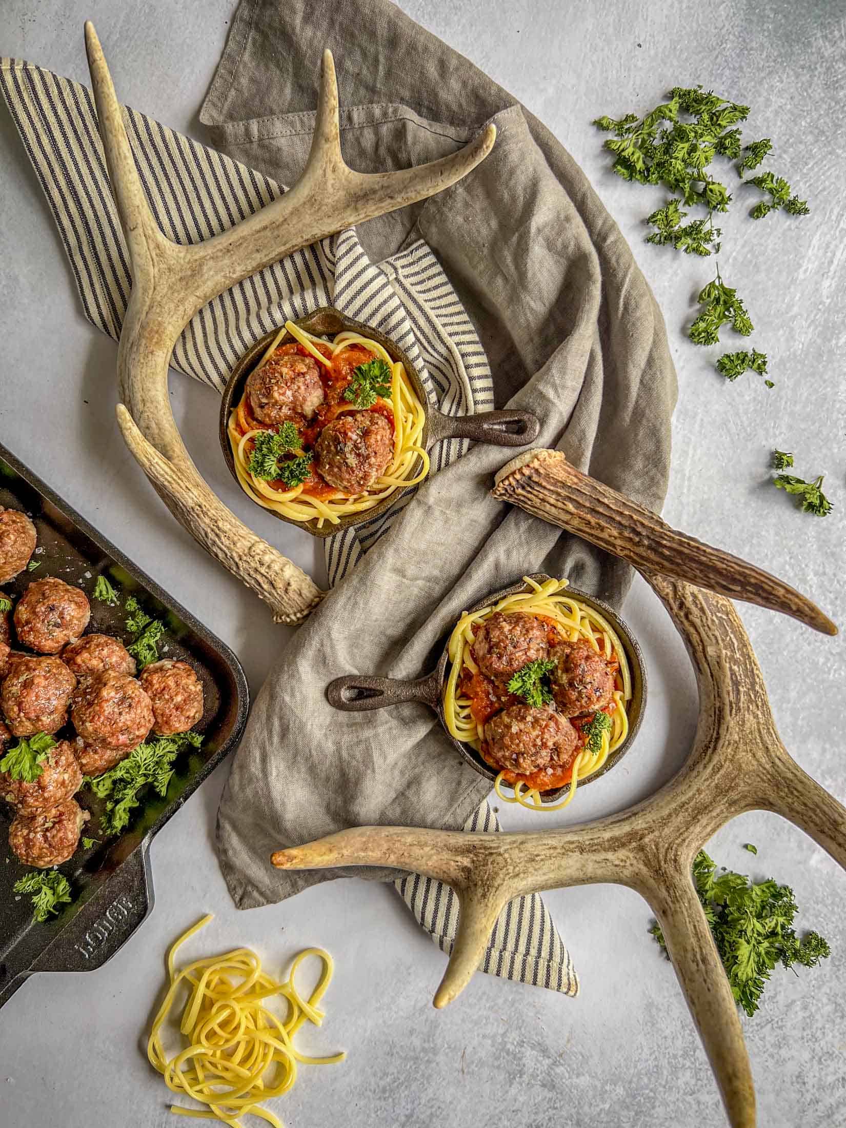 Two mini cast iron skillets filled with  noodles, deer meatballs, and marinara sauce. They have two large deer antlers surrounding them and piles of parsley all around.