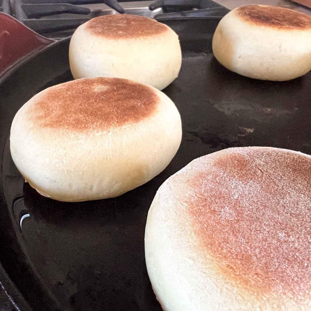 Puffy sourdough english muffins on a cast iron skillet after being flipped once.
