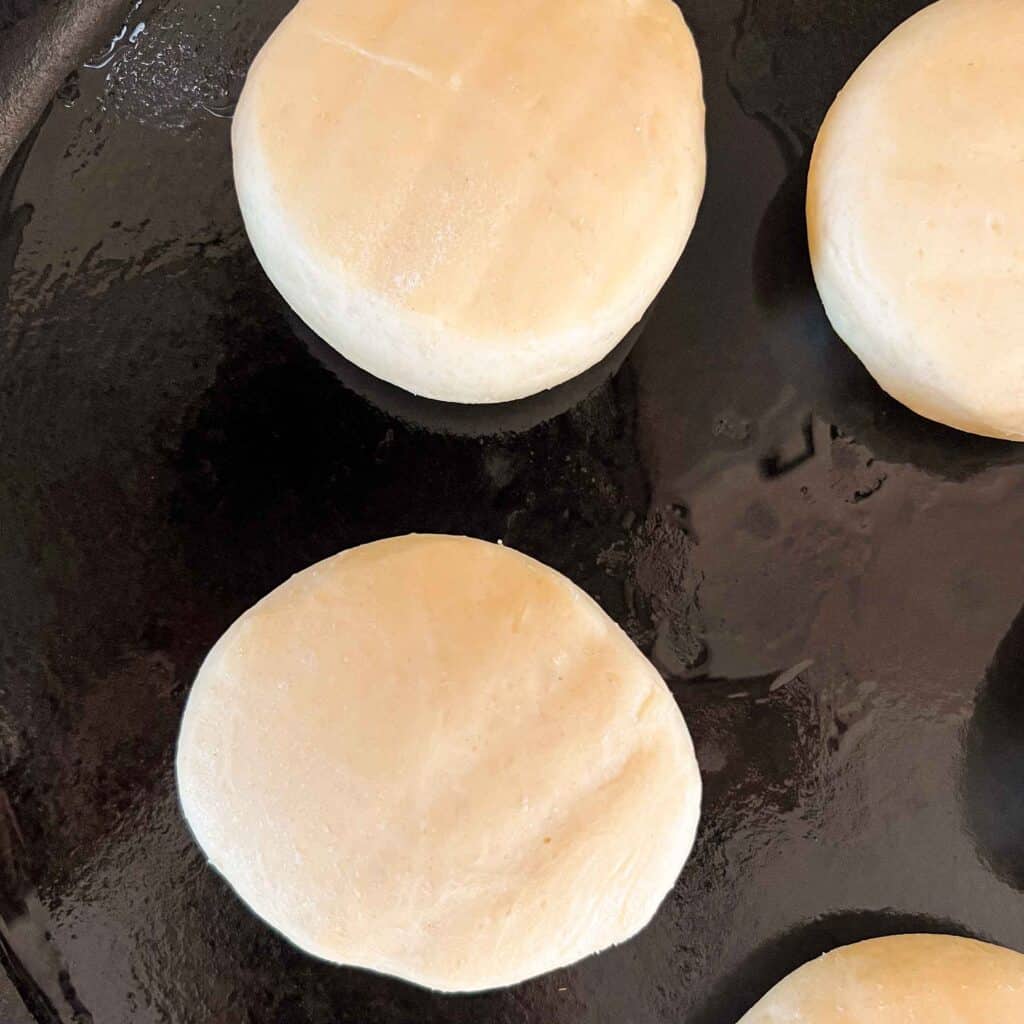 An overhead view of uncooked english muffins on an oiled and preheated cast iron skillet.