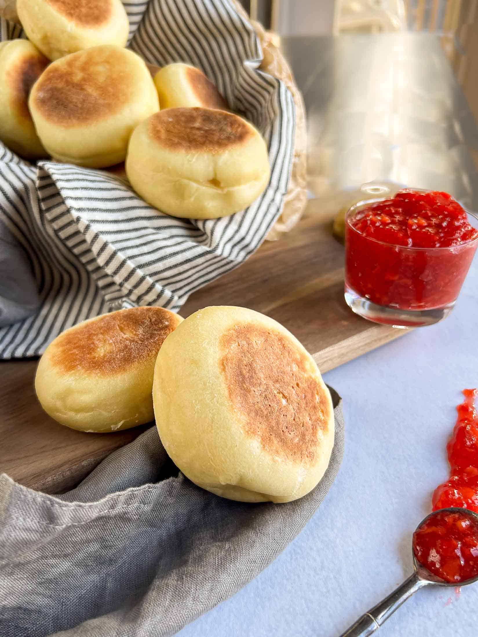 Cooked sourdough english muffins spilling out of a basket with bright strawberry jam beside.