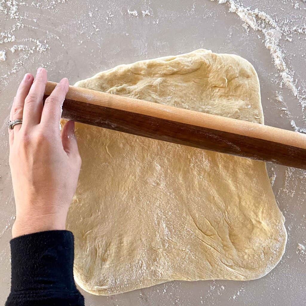 Discard english muffin dough being rolled out on a floured surface with a wooden rolling pin.