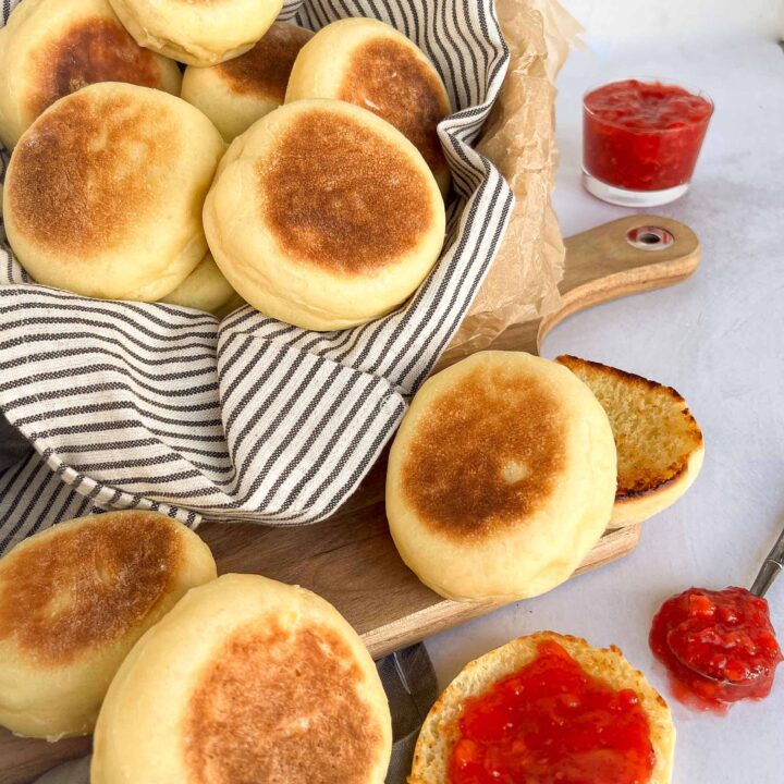 Sourdough discard english muffins spilling out of a linen lines basket with strawberry jam smeared on top of a toasted muffin.